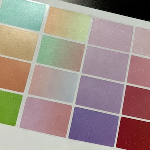 May include: A sheet of rectangular stickers in various pastel colors, including shades of green, orange, pink, purple, and red. Each sticker has a glittery, shimmering finish, creating a textured appearance. The stickers are arranged in a grid pattern.