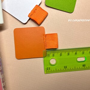 May include: Orange fabric bookmark with a ruler measuring in inches and centimeters. The bookmark is on a light brown surface.