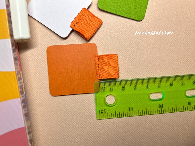 May include: Orange fabric bookmark with a ruler measuring in inches and centimeters. The bookmark is on a light brown surface.
