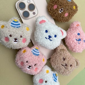 May include: A selection of fluffy bear-shaped phone charms in shades of brown, white, and pink. Each charm has embroidered facial features, with some sporting party hats. A smartphone is partially visible in the background.