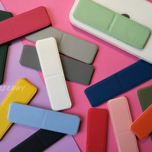 May include: A collection of colorful phone grip stands in various shades, including red, green, blue, and yellow. The rectangular stands are shown on a pink and purple background, with one attached to a white phone. The text "BY CARAFREEPAY" is visible.