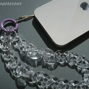 Op de afbeelding: Een transparante plastic kettingtelefoonriem met een paarse karabijnhaak. De riem is bevestigd aan een witte smartphone met een Apple-logo.