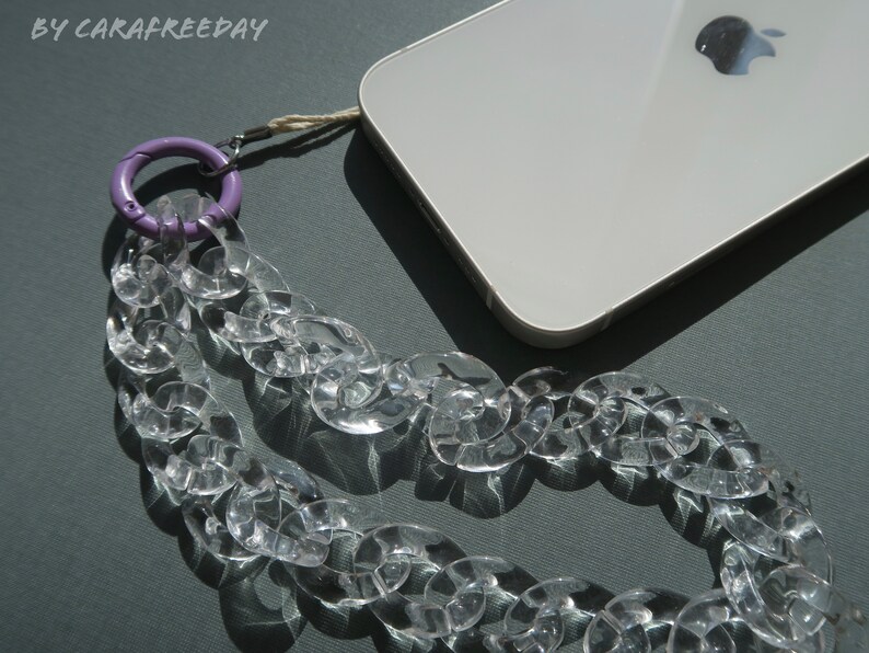 Op de afbeelding: Een transparante plastic kettingtelefoonriem met een paarse karabijnhaak. De riem is bevestigd aan een witte smartphone met een Apple-logo.