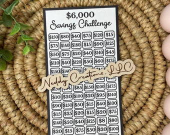 May include: A black and white savings challenge card with the text "$6,000 Savings Challenge." The card features a grid of boxes with various dollar amounts. A wooden cutout of "Neddy Creations, LLC" is placed on the card. The card is on a woven surface.
