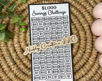 May include: A black and white printable savings challenge chart with the text "$1,000 Savings Challenge" at the top. The chart is filled with dollar amounts ranging from $1 to $20. A wooden sign with the text "Naddy Creations LLC" is placed on top of the chart.