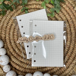May include: Two white gridded paper refill inserts for a planner or notebook. The inserts are packaged in a clear plastic sleeve. A wooden tag with the text "Neddy Creations LLC" is visible in the image.