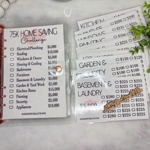 May include: A clear binder with a rose gold spine holds a savings challenge tracker. The tracker lists home improvement categories with amounts. Additional white cards with savings goals are included.