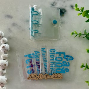 May include: A clear plastic mini three-ring binder with a blue snap closure and matching blue rings. Included are five clear plastic cash envelopes with blue text reading Pets, Alexander, Miscellaneous, Clothing, and Food, with a wooden logo tag.