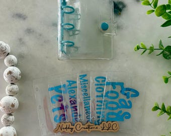 May include: A clear plastic mini three-ring binder with a blue snap closure and matching blue rings. Included are five clear plastic cash envelopes with blue text reading Pets, Alexander, Miscellaneous, Clothing, and Food, with a wooden logo tag.