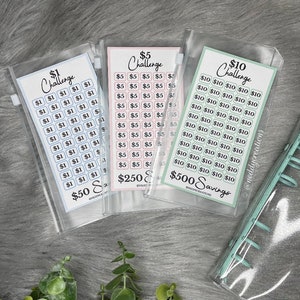 May include: Three clear plastic zippered pouches with printed paper inserts for a savings challenge. The pouches are labeled "$1 Challenge", "$5 Challenge", and "$10 Challenge". The $1 pouch has blue squares with the number 1 printed on them. The $5 pouch has pink squares with the number 5 printed on them. The $10 pouch has green squares with the number 10 printed on them. The pouches are labeled with the total amount of money that can be saved, $50, $250, and $500.