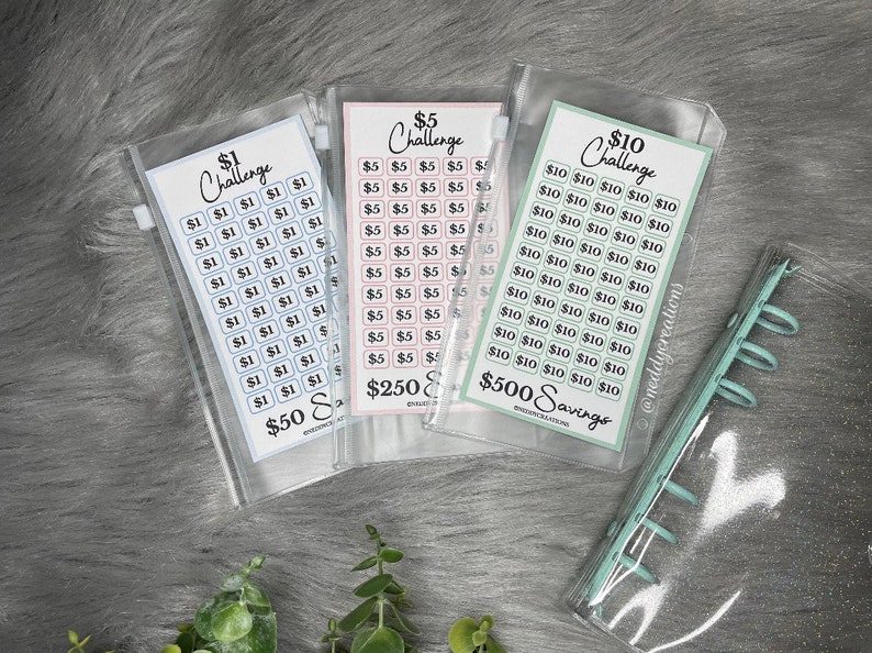 May include: Three clear plastic zippered pouches with printed paper inserts for a savings challenge. The pouches are labeled "$1 Challenge", "$5 Challenge", and "$10 Challenge". The $1 pouch has blue squares with the number 1 printed on them. The $5 pouch has pink squares with the number 5 printed on them. The $10 pouch has green squares with the number 10 printed on them. The pouches are labeled with the total amount of money that can be saved, $50, $250, and $500.