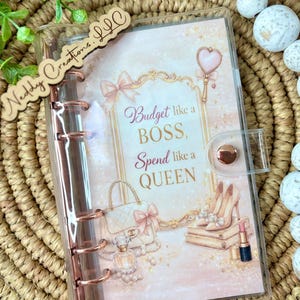 May include: A clear planner with rose gold accents and a decorative insert. The insert features illustrations of a purse, perfume, high heels, and lipstick, with the text "Budget like a BOSS, Spend like a QUEEN."