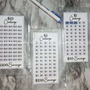 May include: Three clear plastic zippered pouches containing white cards with black text. The cards are labeled with different dollar amounts and the text "Challenge" and "Savings". The cards are designed to help people save money.