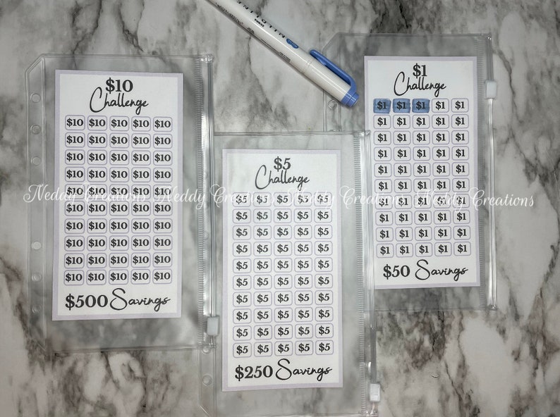 May include: Three clear plastic zippered pouches containing white cards with black text. The cards are labeled with different dollar amounts and the text "Challenge" and "Savings". The cards are designed to help people save money.