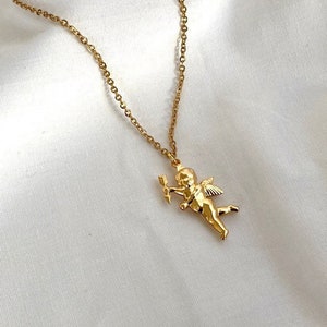 Op de afbeelding: Een gouden ketting met een klein gouden cherubijn hanger die een pijl vasthoudt. De cherubijn heeft vleugels en vliegt.