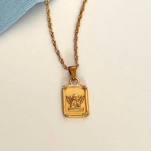 18k gold angel necklace with rectangular cherub pendant on a delicate gold chain displayed on a soft cream background