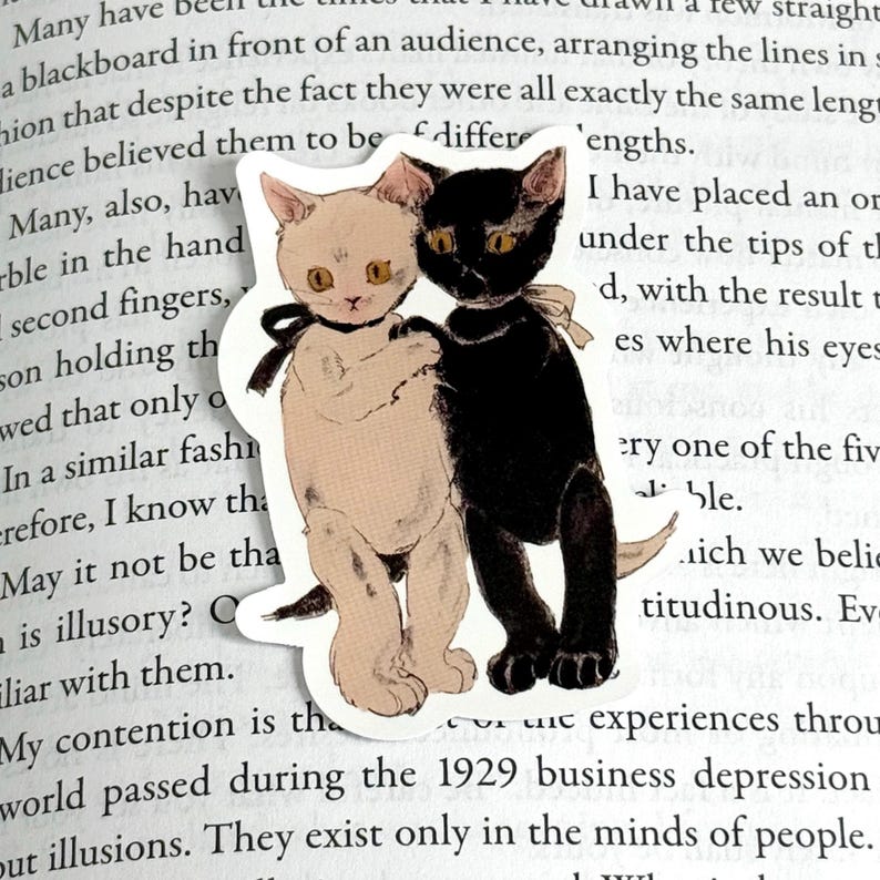 K&ouml;nnte beinhalten: Ein Aufkleber mit einer schwarzen Katze und einer wei&szlig;en Katze mit braunen Augen. Die Katzen umarmen sich. Der Aufkleber befindet sich auf einer Buchseite mit Text.
