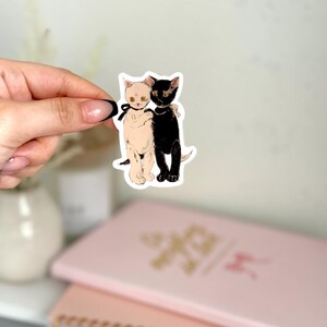 K&ouml;nnte beinhalten: Ein Aufkleber mit einer schwarzen Katze und einer wei&szlig;en Katze, die sich umarmen. Die Katzen haben schwarze Augen und goldene Akzente.