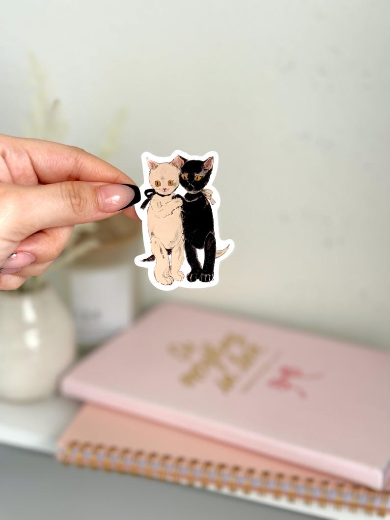 K&ouml;nnte beinhalten: Ein Aufkleber mit einer schwarzen Katze und einer wei&szlig;en Katze, die sich umarmen. Die Katzen haben schwarze Augen und goldene Akzente.