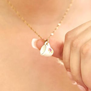 May include: A gold chain necklace with a small white porcelain teacup pendant featuring a pink floral design.