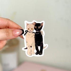 K&ouml;nnte beinhalten: Ein Aufkleber mit einer Illustration von zwei Katzen, einer schwarzen und einer wei&szlig;en. Die Katzen stehen nebeneinander, die wei&szlig;e Katze links und die schwarze Katze rechts. Beide Katzen haben gelbe Augen und tragen schwarze B&auml;nder um den Hals.