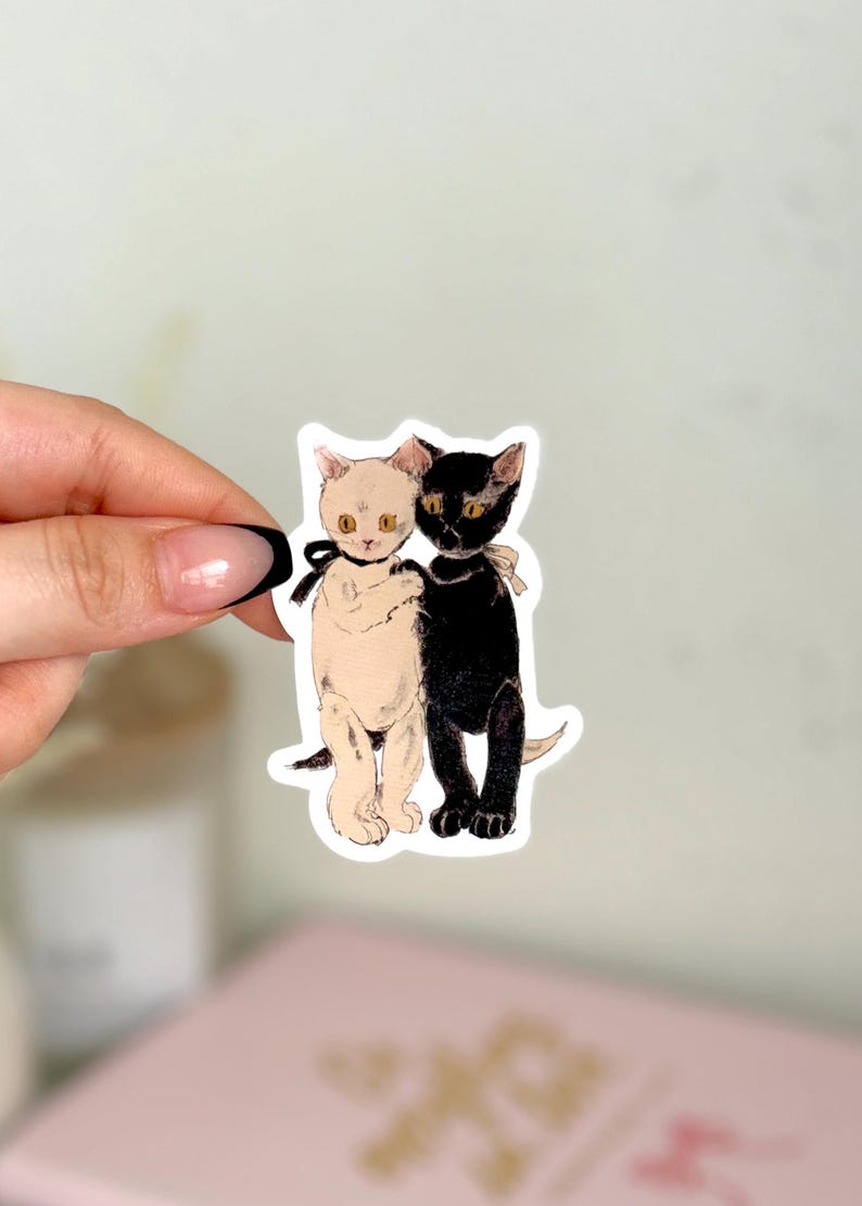K&ouml;nnte beinhalten: Ein Aufkleber mit einer Illustration von zwei Katzen, einer schwarzen und einer wei&szlig;en. Die Katzen stehen nebeneinander, die wei&szlig;e Katze links und die schwarze Katze rechts. Beide Katzen haben gelbe Augen und tragen schwarze B&auml;nder um den Hals.