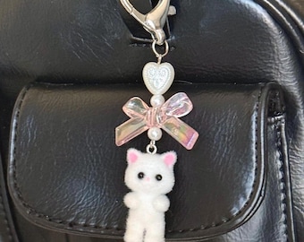 May include: A white plush cat charm with pink bows and pearl beads hanging from a silver and gold chain. The charm is attached to a black leather bag.