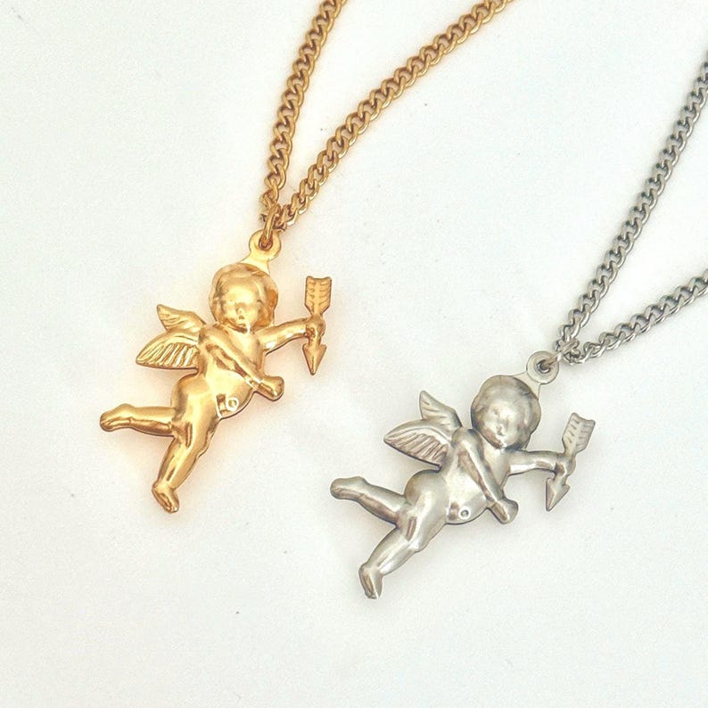 Op de afbeelding: Twee kettingen met engel-cupido hangers. De ene ketting heeft een gouden hanger en de andere een zilveren hanger. Beide hangers tonen een cupido die een pijl vasthoudt.