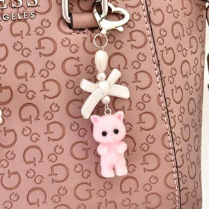Op de afbeelding: Een roze pluche kattenhanger met een witte strik en parel accenten hangt aan een zilveren sleutelhanger. De hanger is bevestigd aan een roze handtas met een herhalend "G" patroon.