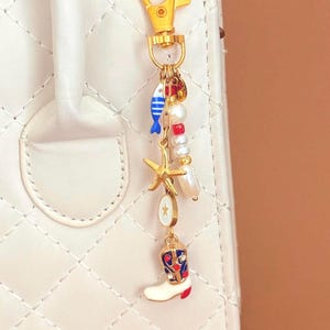 May include: A gold-colored keychain with various charms, including a blue fish, a starfish, and a white cowboy boot with red and blue accents. The keychain is attached to a white quilted handbag.