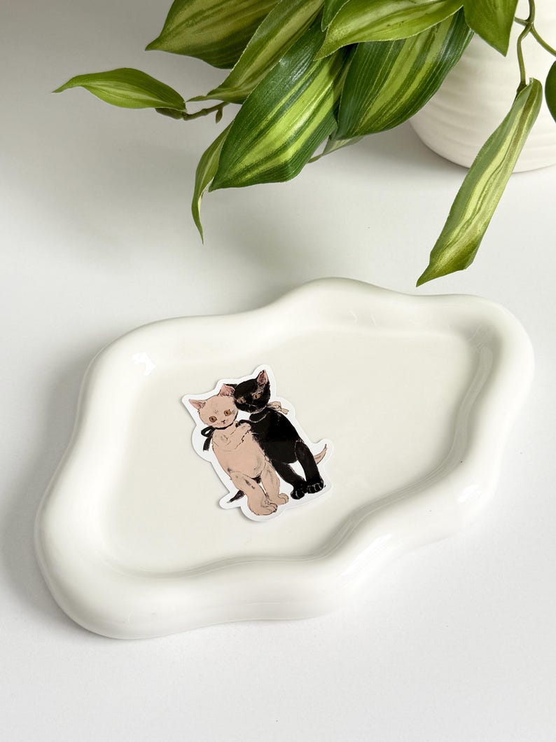 K&ouml;nnte beinhalten: Eine wei&szlig;e Keramikschale mit welligem Rand, auf der ein Aufkleber mit zwei sich umarmenden Katzen angebracht ist. Die Katze links ist wei&szlig; mit rosa Ohren und einer schwarzen Schleife, w&auml;hrend die Katze rechts schwarz mit einer wei&szlig;en Schleife ist.