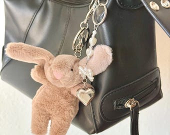 May include: A black leather handbag with a brown plush bunny keychain. The keychain has a silver heart charm and a silver chain with white beads.