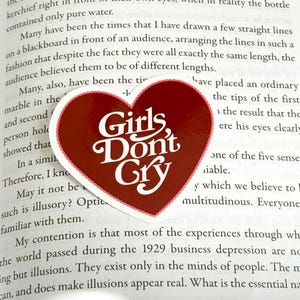Puede incluir: Un marcapáginas en forma de corazón rojo con el texto "Girls, Don't Cry" en letras blancas. El marcapáginas está encima de un libro.