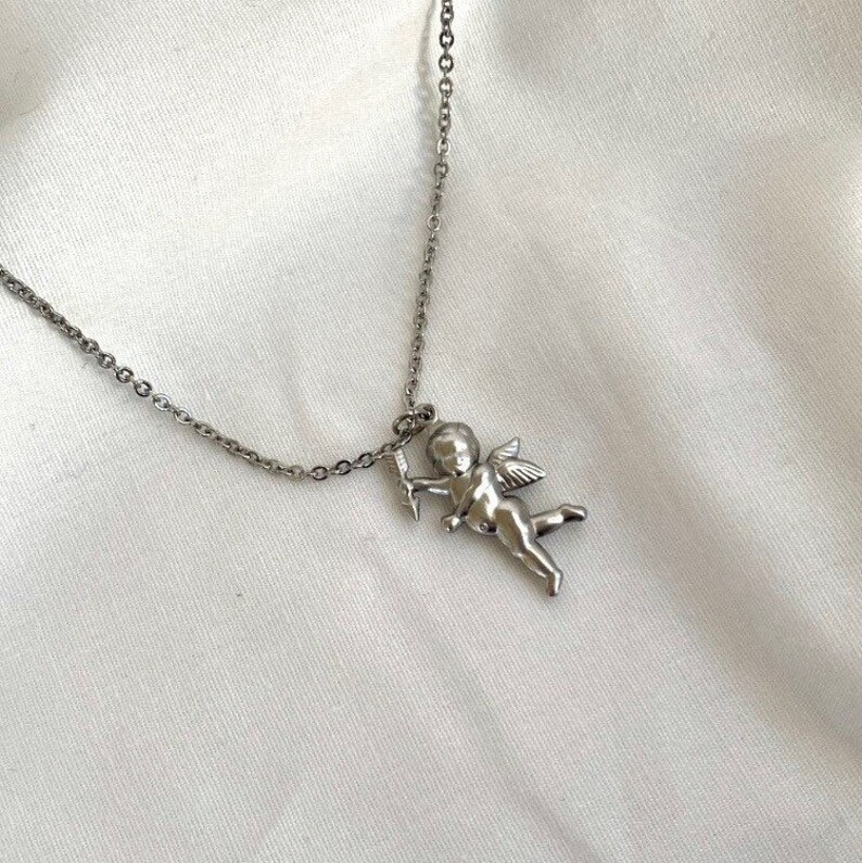 Op de afbeelding: Een zilveren ketting met een zilveren cherubijn hanger die een pijl vasthoudt. De hanger is gedetailleerd en heeft vleugels.