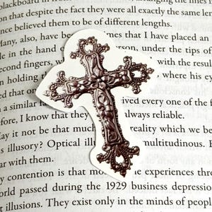 Könnte beinhalten: Ein silberfarbenes Metall-Kreuz Lesezeichen mit filigranen Blumendesigns. Das Lesezeichen befindet sich auf einer Seite eines Buches mit Text.