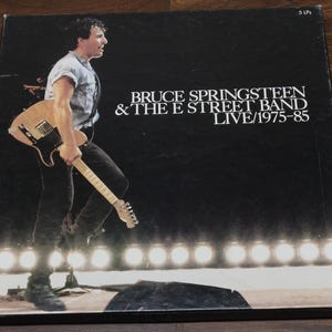 May include: A black vinyl record album cover featuring Bruce Springsteen and the E Street Band. The cover shows a black and white photo of Bruce Springsteen playing a guitar on stage. The text on the cover reads "Bruce Springsteen & The E Street Band LIVE/1975-85".