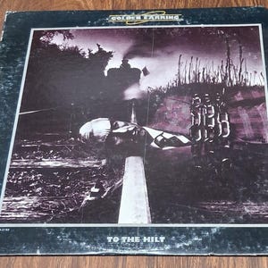 May include: A square vinyl record jacket featuring the Golden Earring album To The Hilt. The cover art displays a monochromatic purple-toned scene of a train on tracks with a large hand in the foreground. The text Golden Earring and To The Hilt are printed on the front.