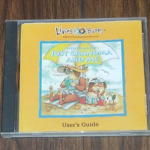 May include: A CD case with a yellow cover featuring the title "Just Grandma and Me" by Mercer Mayer. The cover art depicts cartoon characters on a beach under an umbrella. The text "User's Guide" is at the bottom.