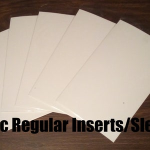 May include: Five white comic book inserts or sleeves, also known as comic book bags, are stacked on a wooden surface. The text "Comic Regular Inserts/Sleeves" is visible at the bottom of the image.