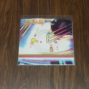 May include: A Def Leppard album cover in a protective sleeve. The album art features the band's logo in yellow and red, with a blue and white background. The text "Pour Some Sugar On Me" is visible.
