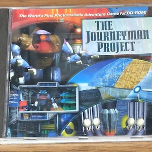 May include: CD-ROM case for the computer game The Journeyman Project. The cover art features a futuristic, sci-fi scene with a robot and a large, domed structure.