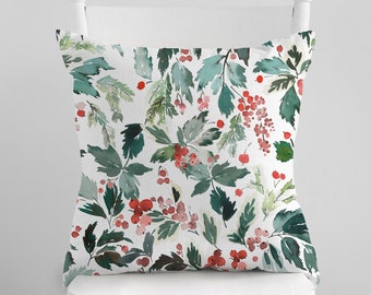 Pode incluir: Almofada quadrada com um design em aquarela de folhas verdes e bagas vermelhas sobre um fundo branco. A almofada está colocada numa cadeira branca, exibindo um padrão festivo inspirado na natureza.