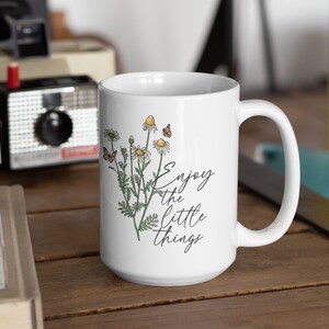 May include: White ceramic mug with a floral design featuring daisies and butterflies, with the text "Enjoy the little things" in a script font. The mug has a large handle and is suitable for hot or cold beverages.