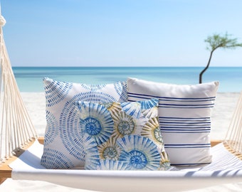 May include: Three decorative throw cushions on a white hammock swing. The cushions feature blue and white seashell and striped patterns. The hammock is suspended by white rope, set against a beach and ocean backdrop.