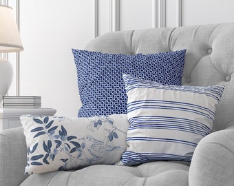 May include: A gray upholstered armchair with three decorative pillows in blue and white patterns. A white side table with a lamp and vase of flowers sits beside the chair. The room has white paneling.