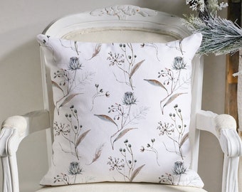 May include: A square white throw pillow with a botanical pattern of muted blue, brown, and green floral elements. The pillow is placed on a white antique chair, suggesting a home decor theme.