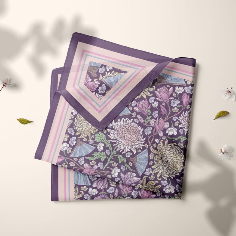 May include: A folded square scarf with a floral pattern in shades of purple, pink, and green. The design includes chrysanthemums, fans, and other floral elements. The scarf has a purple border with a light pink and blue inner border.
