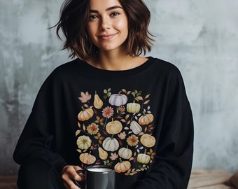 Könnte beinhalten: Schwarzes Sweatshirt mit einem Aquarelldesign aus Kürbissen, Birnen und Herbstlaub in Orange-, Weiß- und Gelbtönen. Die Person auf dem Bild hält einen dunkelgrauen Becher.