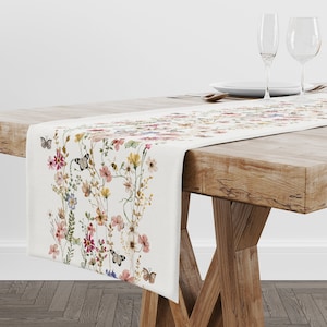 Op de afbeelding: Crèmekleurige tafelloper met een aquarel bloemenontwerp en vlinders. De loper toont kleurrijke bloemen in roze, geel en blauw. Hij ligt op een houten tafel.