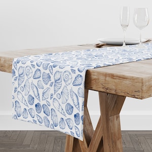May include: A white table runner with a blue seashell pattern. The runner is laid on a wooden table with two wine glasses and a plate.